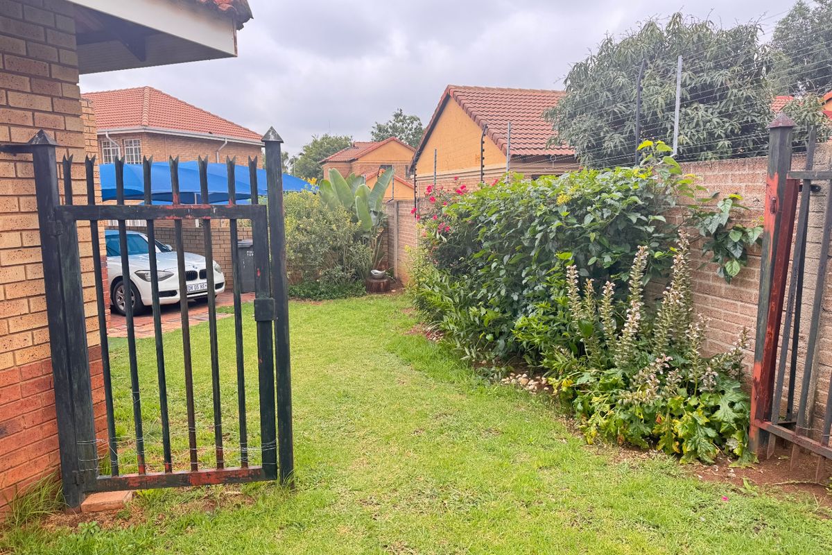 A private rear garden area featuring a lawn, established perimeter landscaping, and a secure metal gate with electric fencing along the boundary wall.