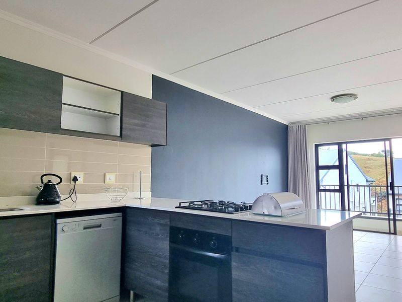 Modern open-plan kitchen featuring dark wood cabinetry, gas hob, built-in oven, and tiled flooring, flowing seamlessly into a bright living area with balcony access.