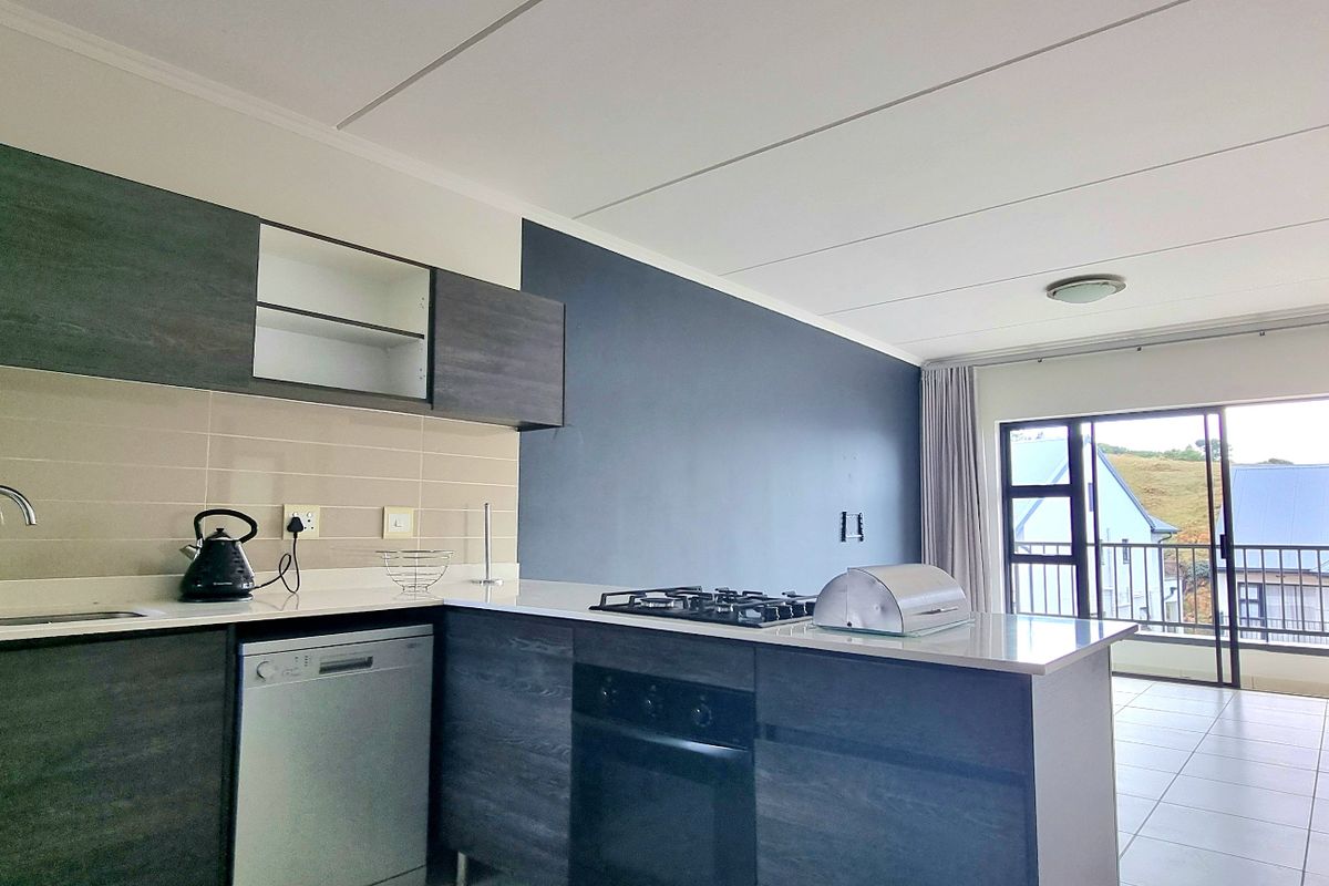 Modern open-plan kitchen featuring dark wood cabinetry, gas hob, built-in oven, and tiled flooring, flowing seamlessly into a bright living area with balcony access.