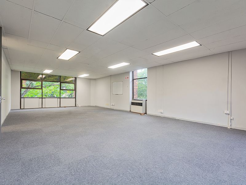 A spacious, empty commercial office featuring a suspended ceiling with recessed lighting, carpeted flooring, and large windows providing natural light.