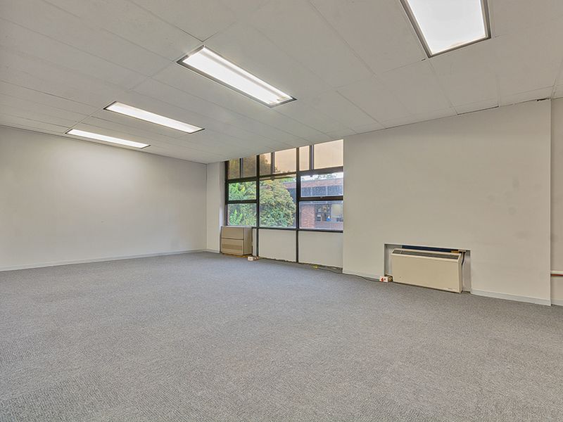 A vacant commercial office space featuring neutral carpeting, a suspended ceiling with recessed lighting, and a wall-mounted HVAC unit.