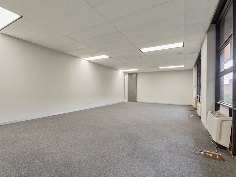 A spacious, empty commercial office featuring carpeted flooring, a suspended ceiling with recessed lighting, and large windows with an integrated HVAC unit.