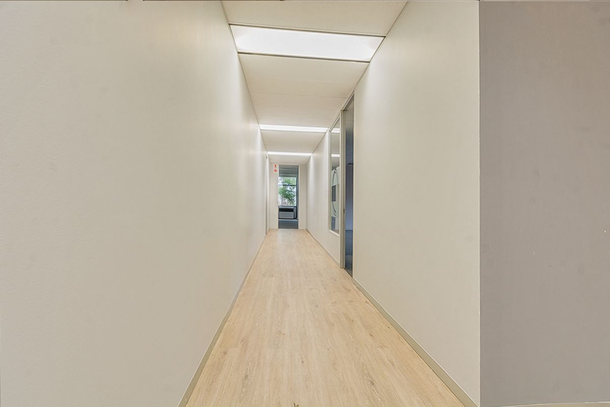 A clean, well-lit commercial hallway featuring wood-look vinyl flooring and recessed ceiling lighting, providing access to adjacent office spaces.