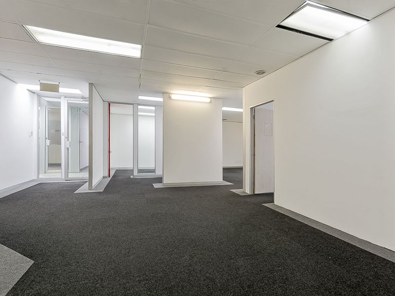An open-plan commercial office space featuring neutral walls, dark carpet tiles, suspended ceilings with recessed lighting, and glass partition walls.