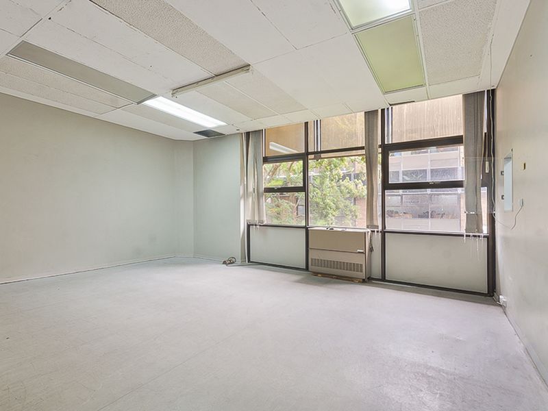 An empty commercial office space featuring large windows, a suspended ceiling with integrated lighting, and a wall-mounted HVAC unit.