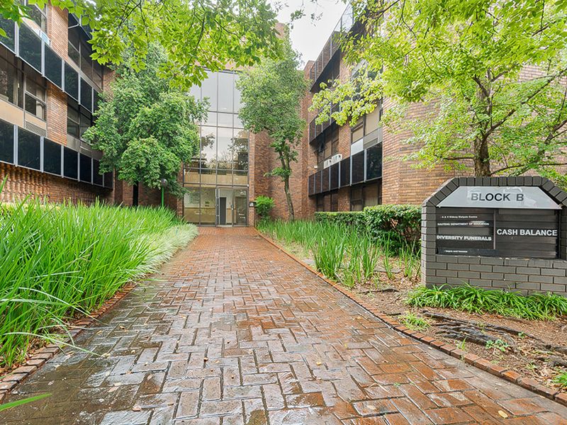 Front entrance view of Block B, featuring a brick facade, glass curtain wall, and a paved walkway leading to the main building access.
