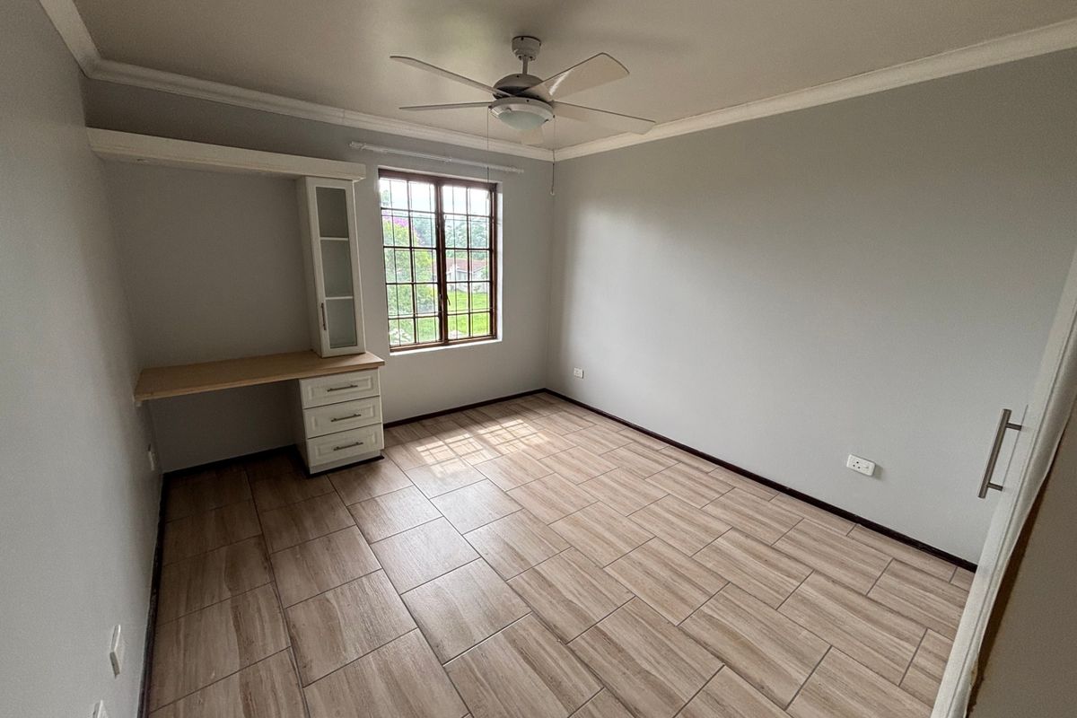 A bright, empty bedroom featuring modern wood-look tiled flooring, a built-in desk with storage, and a ceiling fan.