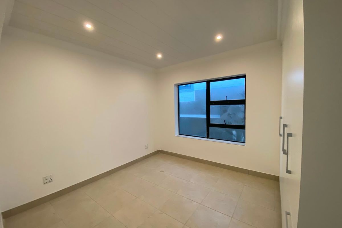 A clean, empty bedroom featuring neutral tiled flooring, built-in cupboards, recessed downlighting, and a modern aluminium-framed window.