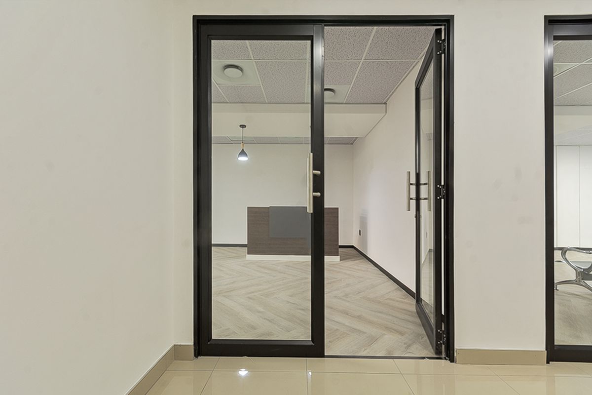 A modern, professional office space featuring black aluminium glass doors, herringbone vinyl flooring, and a built-in reception desk under a suspended ceiling.