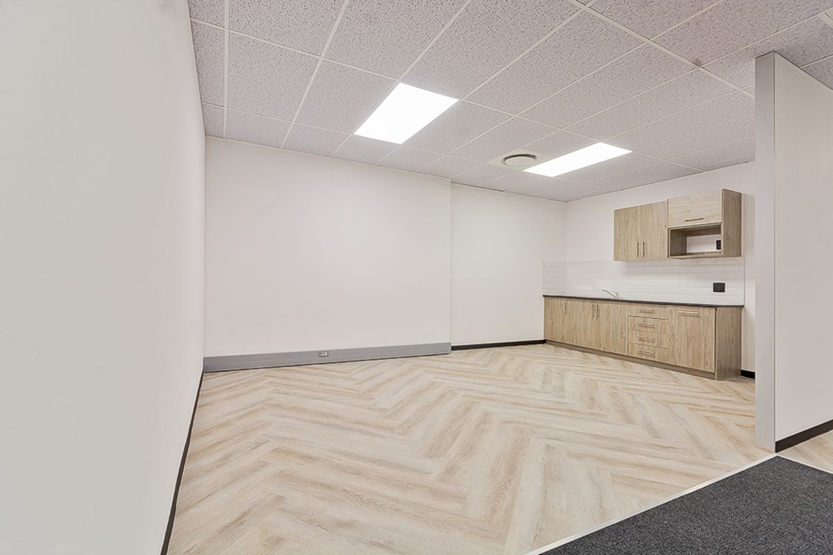 A clean, modern commercial kitchenette featuring light wood-grain cabinetry, herringbone vinyl flooring, and a neutral tiled backsplash in an open-plan office space.