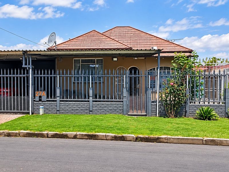 Front elevation of a residential property featuring a tiled roof, secure metal perimeter fencing, a paved driveway, and a covered carport area.