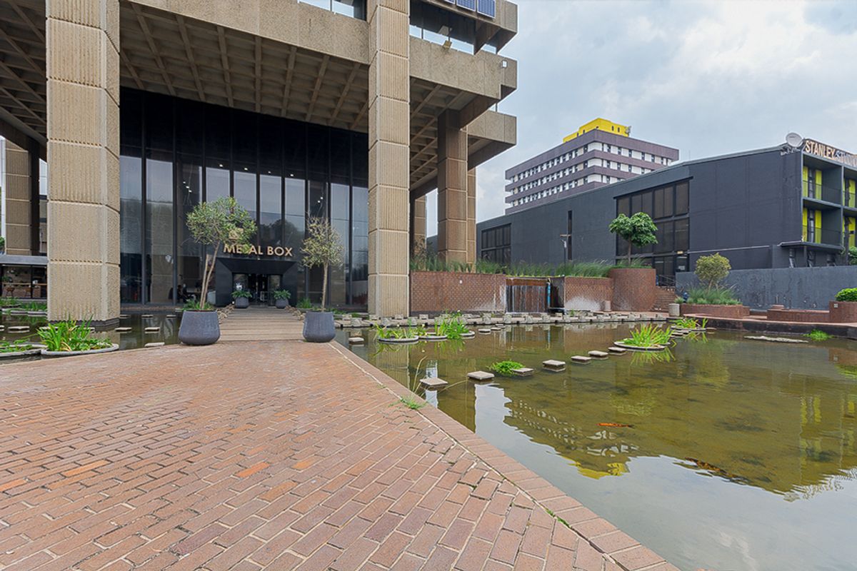 Front entrance of the Metal Box commercial building featuring a prominent concrete structure, glass facade, and a decorative water feature with brick paving.