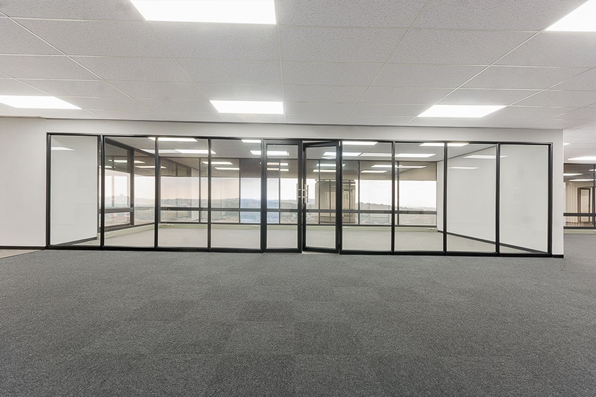 A modern, open-plan commercial office space featuring glass partitioning, suspended ceilings with recessed lighting, and neutral carpeting.