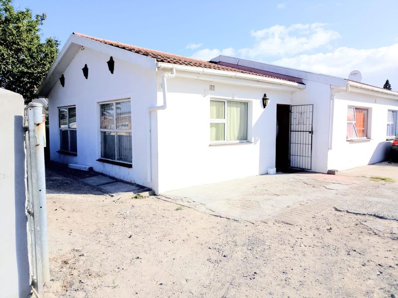 Front exterior view of a single-story house featuring white painted walls, tiled roof, security gate, and an unpaved driveway area.