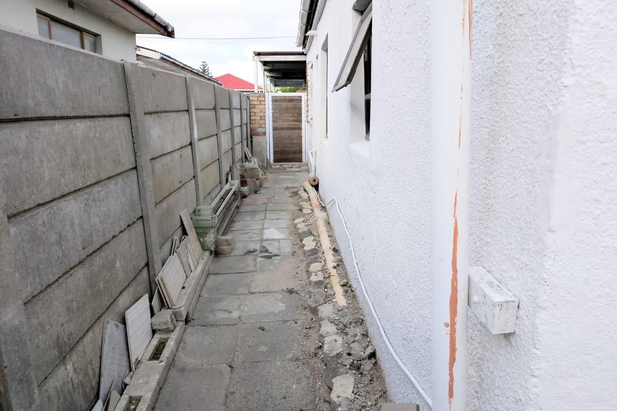 A narrow side passage of the house featuring a concrete boundary wall, paved walkway, and exposed exterior wall sections requiring maintenance.