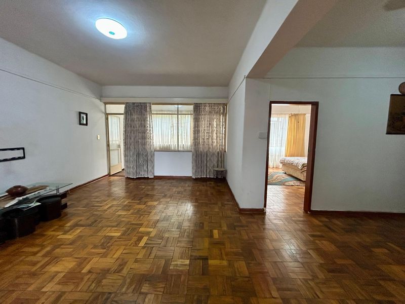 Spacious living area featuring original parquet flooring and neutral walls, offering a versatile layout with direct access to an adjoining bedroom.