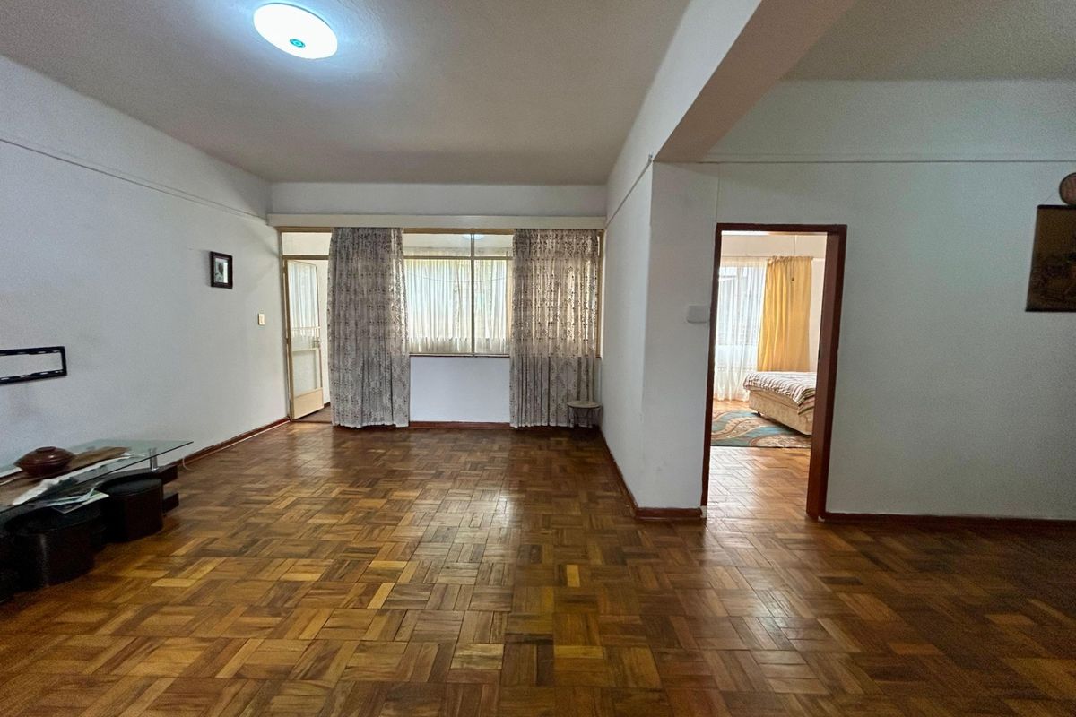 Spacious living area featuring original parquet flooring and neutral walls, offering a versatile layout with direct access to an adjoining bedroom.