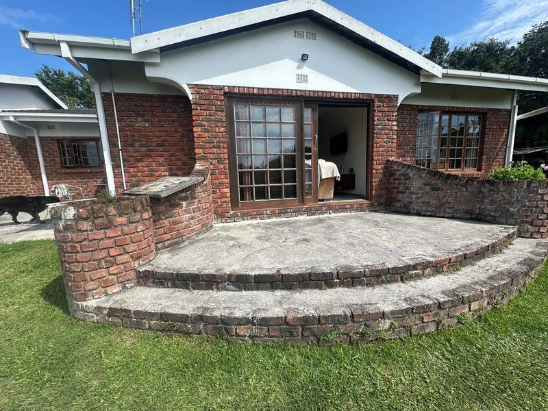 Rear view of a brick house featuring a raised concrete patio, sliding glass door access, and a surrounding garden area.