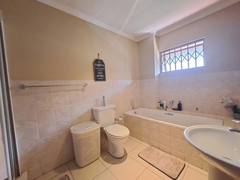 A functional bathroom featuring a standard bathtub, pedestal basin, and toilet with tiled walls and flooring, complemented by a security-barred window.