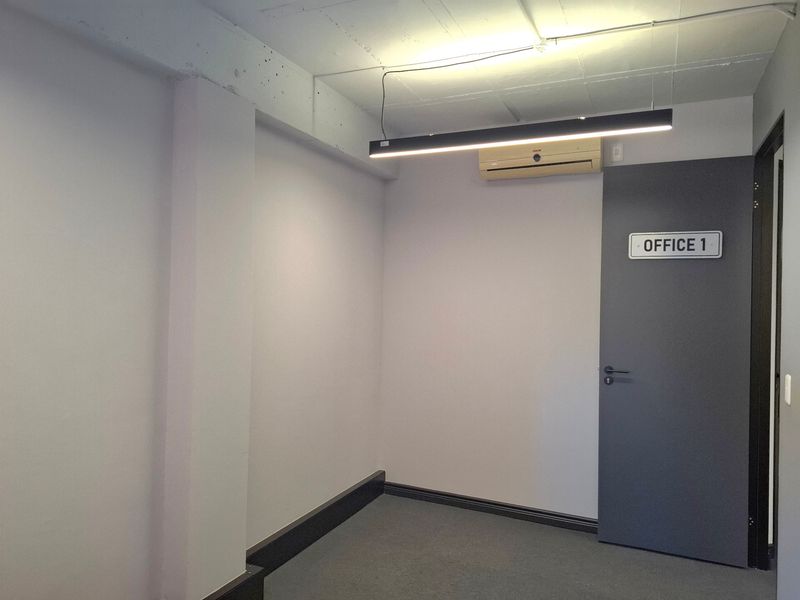 A clean, minimalist office space featuring exposed concrete ceilings, modern linear lighting, a wall-mounted air conditioning unit, and neutral carpet flooring.