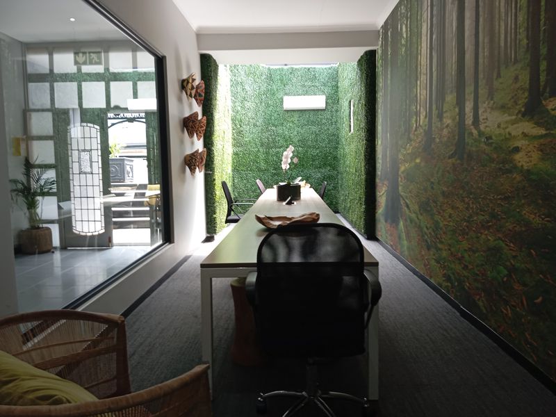 Modern office space featuring a long meeting table, artificial grass wall accents, and a large glass partition overlooking an adjacent area.