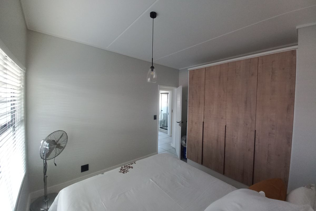 A well-maintained bedroom featuring modern wood-finish built-in wardrobes, a contemporary pendant light, and venetian blinds, with an adjoining en-suite bathroom visible.