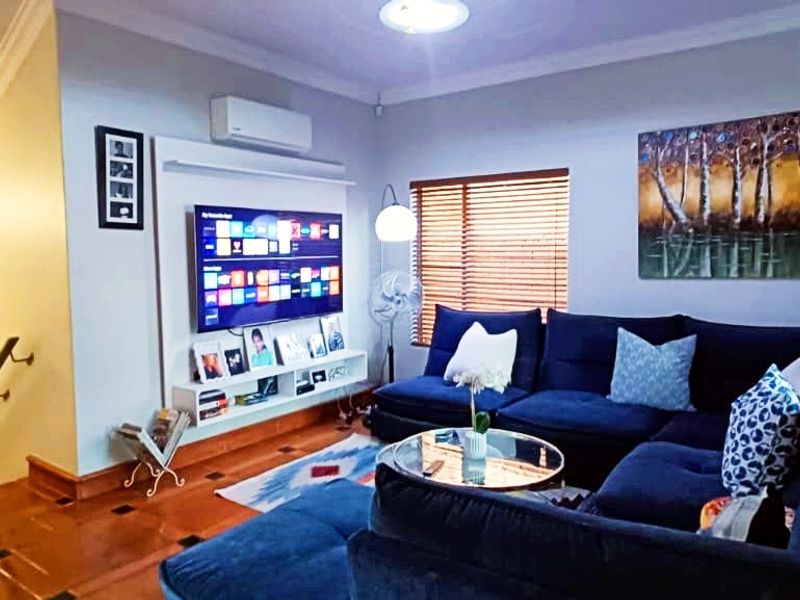 A comfortable living room featuring polished wooden flooring, timber blinds, and a wall-mounted air conditioning unit, with an adjacent hallway and staircase.