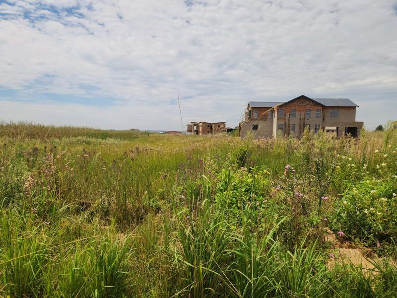 A view of a vacant residential plot featuring tall, overgrown grass with partially constructed buildings visible in the background.