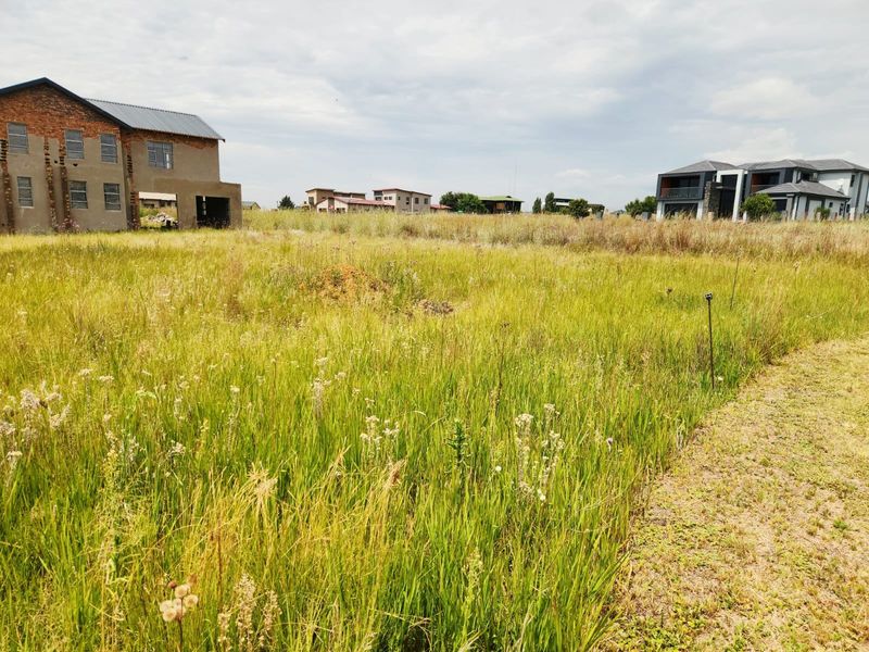 A view of a vacant residential plot featuring tall grass, with neighboring houses visible in the background.
