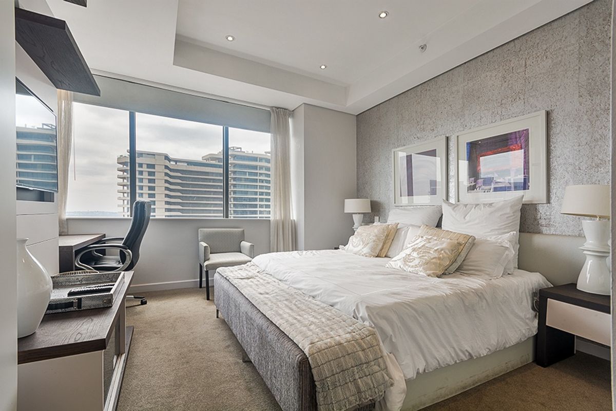 A modern, well-lit bedroom featuring a textured feature wall, large windows with city views, and integrated desk space.