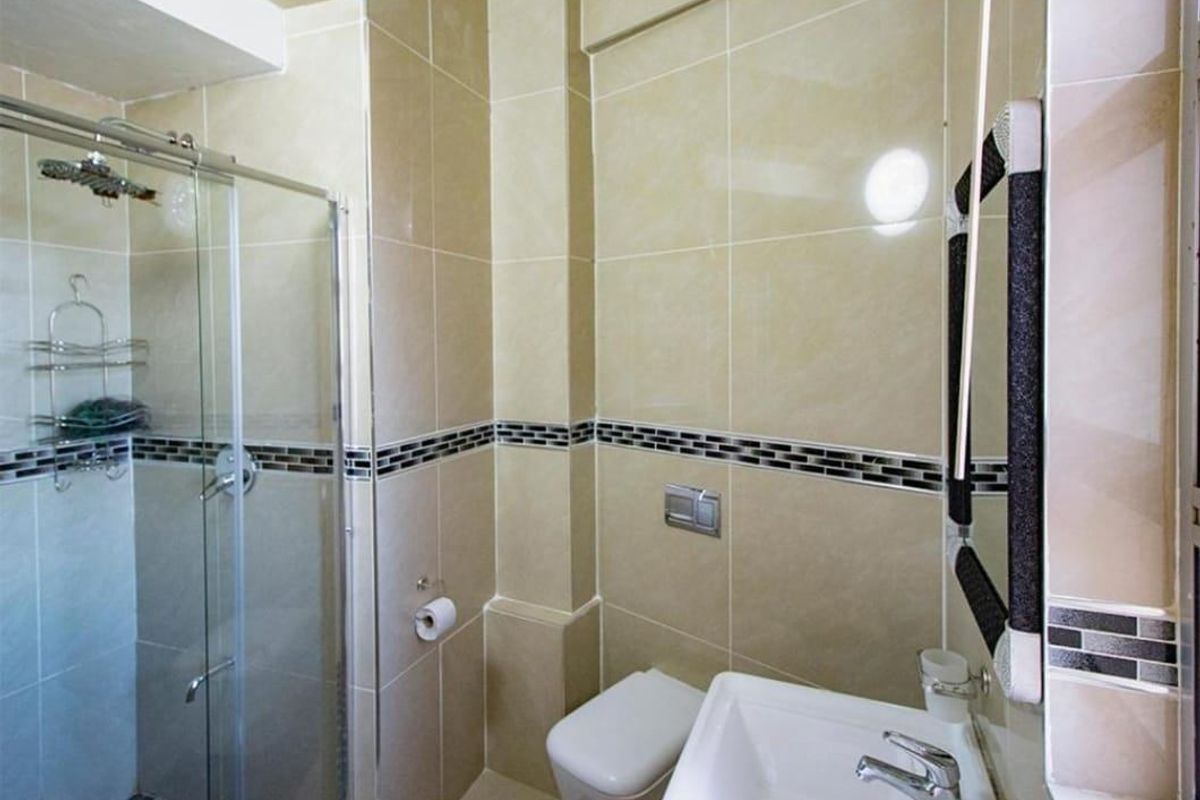 A compact, modern bathroom featuring a glass-enclosed shower, wall-mounted toilet, and neutral ceramic tiling with a decorative mosaic border.