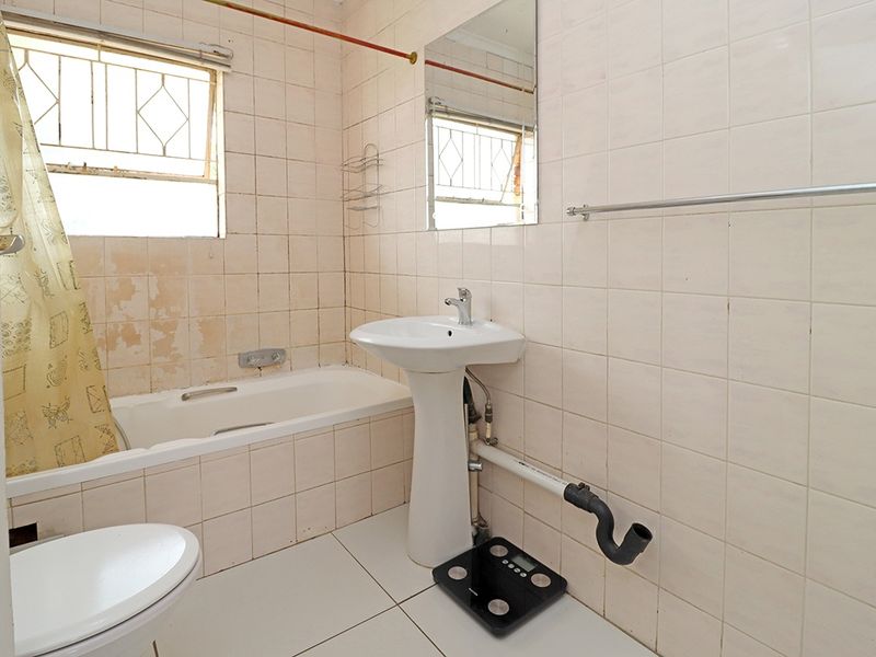 A dated bathroom featuring a pedestal basin, bathtub, and toilet, with visible exposed plumbing and worn wall tiling requiring renovation.