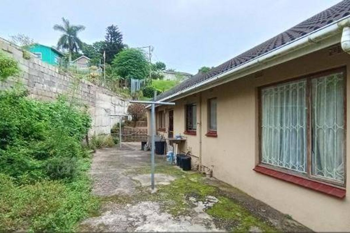 Rear exterior view of the property featuring a concrete paved area, retaining wall, and established garden space requiring maintenance.