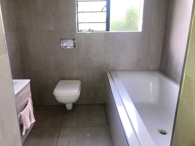 A modern bathroom featuring grey tiled walls and flooring, a wall-mounted toilet with a concealed cistern, and a built-in bathtub.