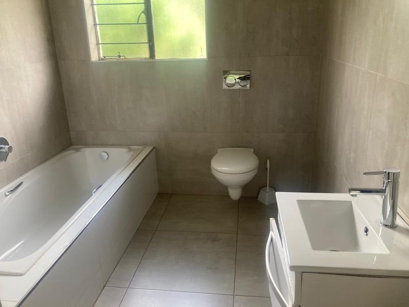A modern, neutral-toned bathroom featuring a wall-mounted toilet, a built-in bathtub, and a vanity basin with contemporary chrome fixtures.