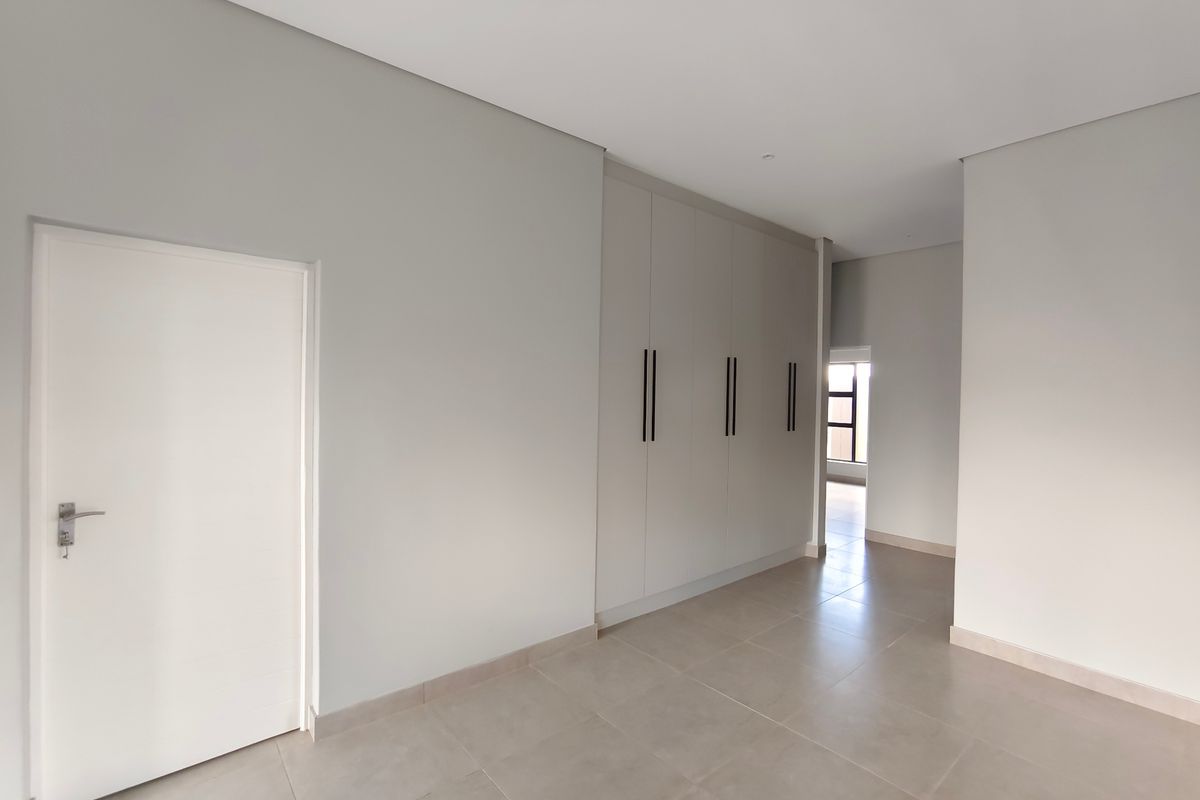 A bright, modern, and empty bedroom featuring neutral tiled flooring, clean white walls, and sleek built-in cupboards with contemporary black handles.