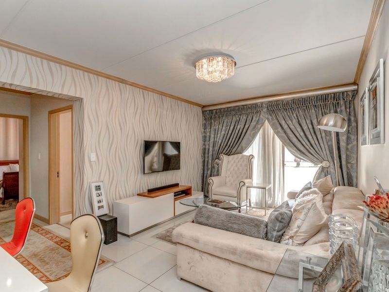 A well-maintained living room featuring tiled flooring, decorative wallpaper, and a crystal ceiling light fixture, with direct access to an adjacent bedroom.