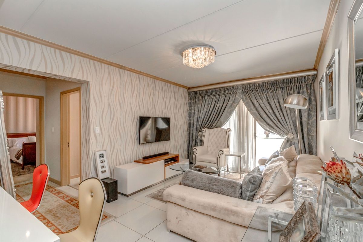 A well-maintained living room featuring tiled flooring, decorative wallpaper, and a crystal ceiling light fixture, with direct access to an adjacent bedroom.