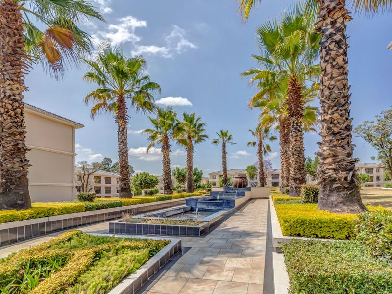 A well-maintained communal garden area featuring a central water fountain, paved walkways, and mature palm trees within an apartment complex.