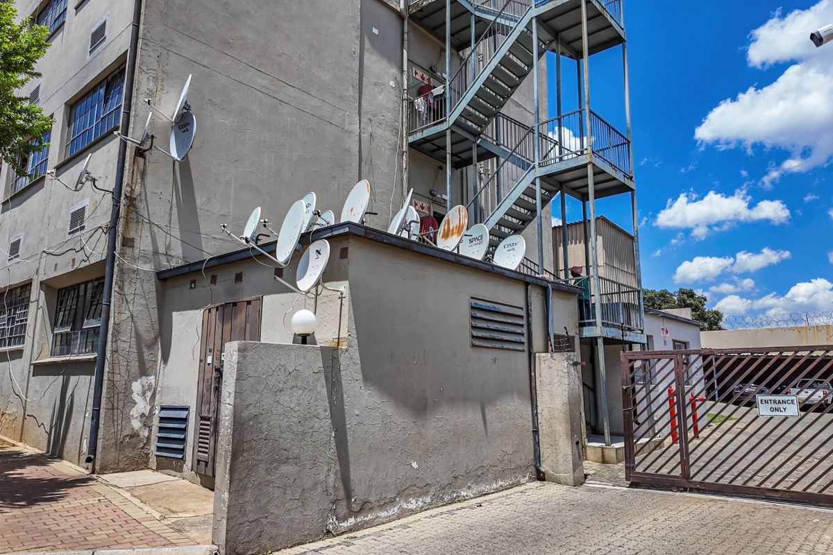 Rear exterior view of the apartment building featuring a metal fire escape staircase, multiple satellite dishes, and a gated entrance.