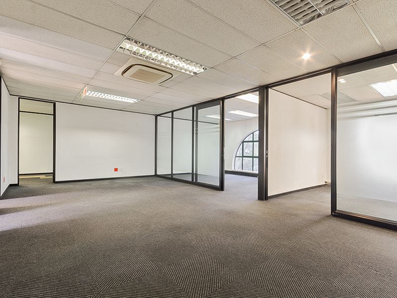 Spacious open-plan commercial office area featuring glass partitions, suspended ceilings with integrated lighting, ducted air conditioning, and carpeted flooring.