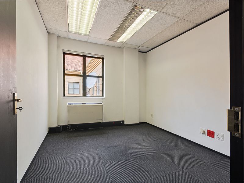 A vacant commercial office space featuring carpeted flooring, a suspended ceiling with integrated lighting, and a wall-mounted HVAC unit.