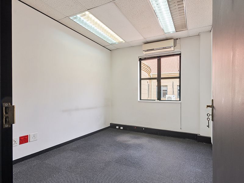 A vacant, compact office space featuring carpeted flooring, a suspended ceiling with integrated lighting, and a wall-mounted air conditioning unit.