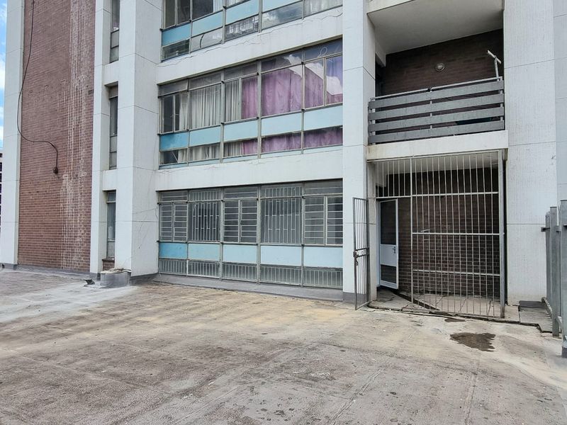 Rear exterior view of an apartment building featuring a concrete courtyard, security gate access, and multi-level balconies with metal window frames.