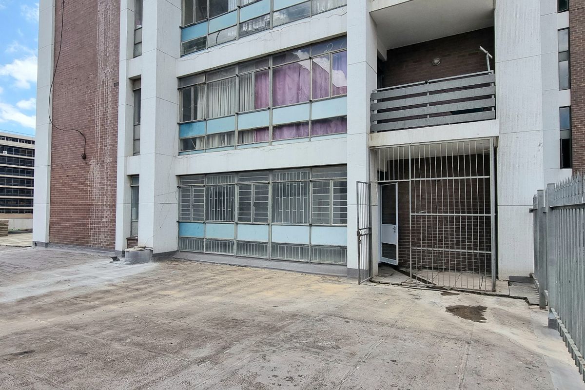 Rear exterior view of an apartment building featuring a concrete courtyard, security gate access, and multi-level balconies with metal window frames.