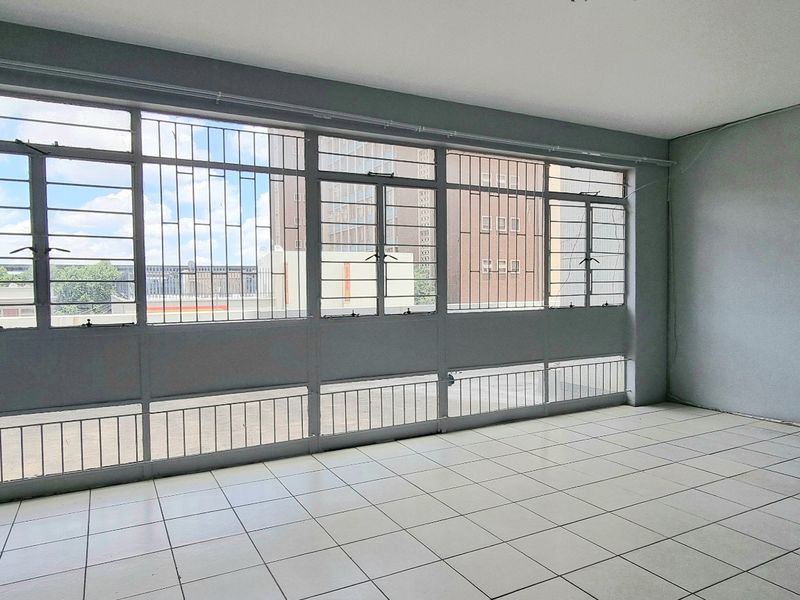 An empty, sunlit living area featuring large metal-framed windows with security bars and neutral tiled flooring, offering a blank canvas for renovation.