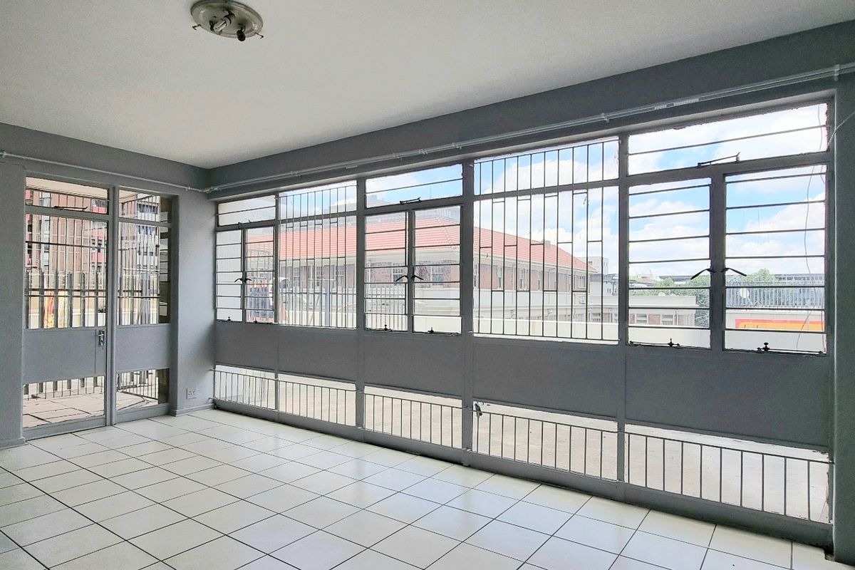 An empty, sunlit living area featuring tiled flooring, large metal-framed windows with security bars, and direct access to an outdoor space.