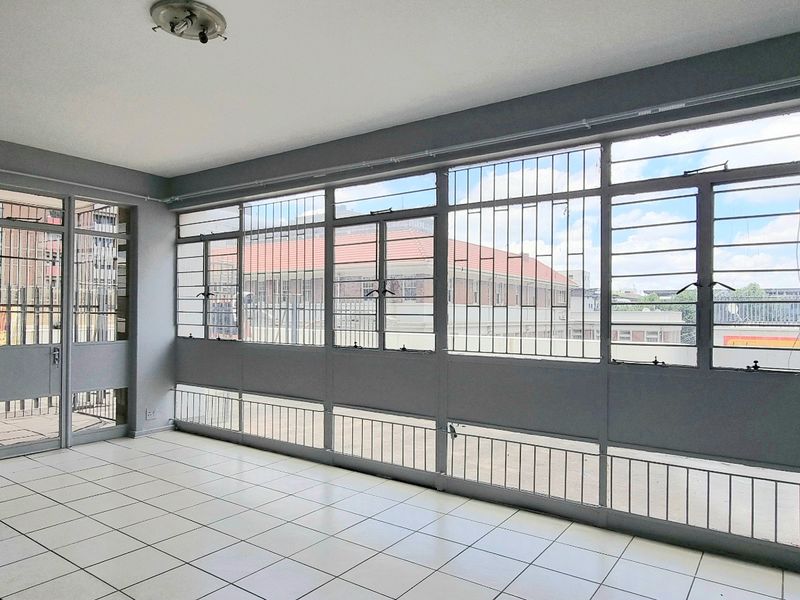 An empty, sunlit living area featuring tiled flooring, large metal-framed windows with security bars, and direct access to an outdoor space.