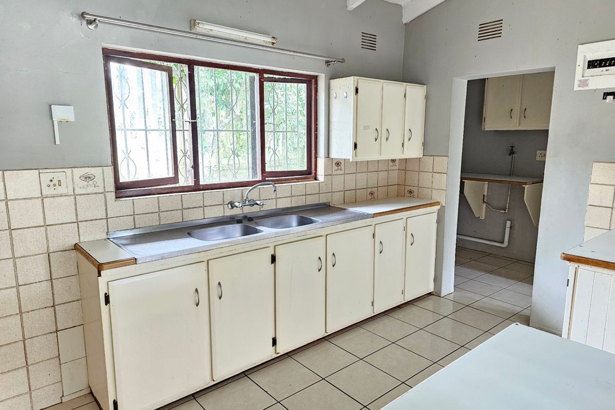 A dated kitchen featuring built-in cabinetry, a double basin sink, and tiled splashbacks, with an adjacent scullery area and exposed beam ceiling.
