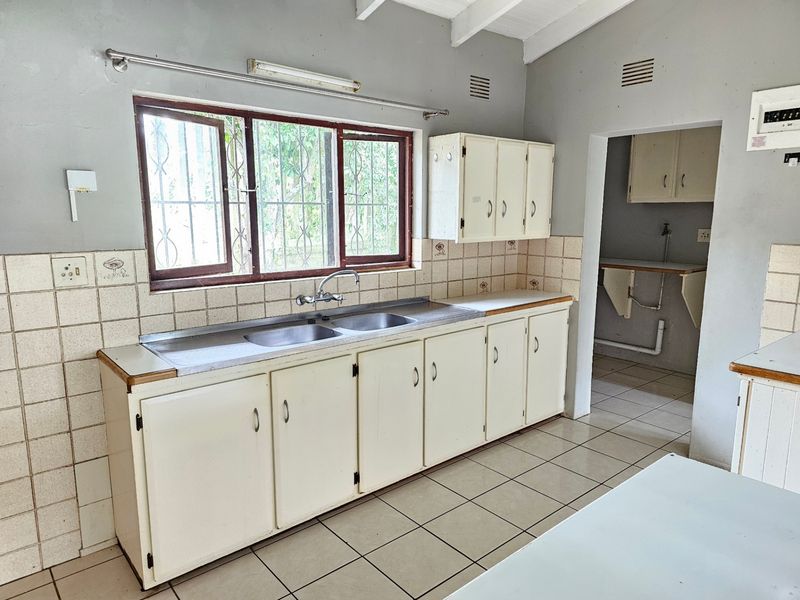 A dated kitchen featuring built-in cabinetry, a double basin sink, and tiled splashbacks, with an adjacent scullery area and exposed beam ceiling.